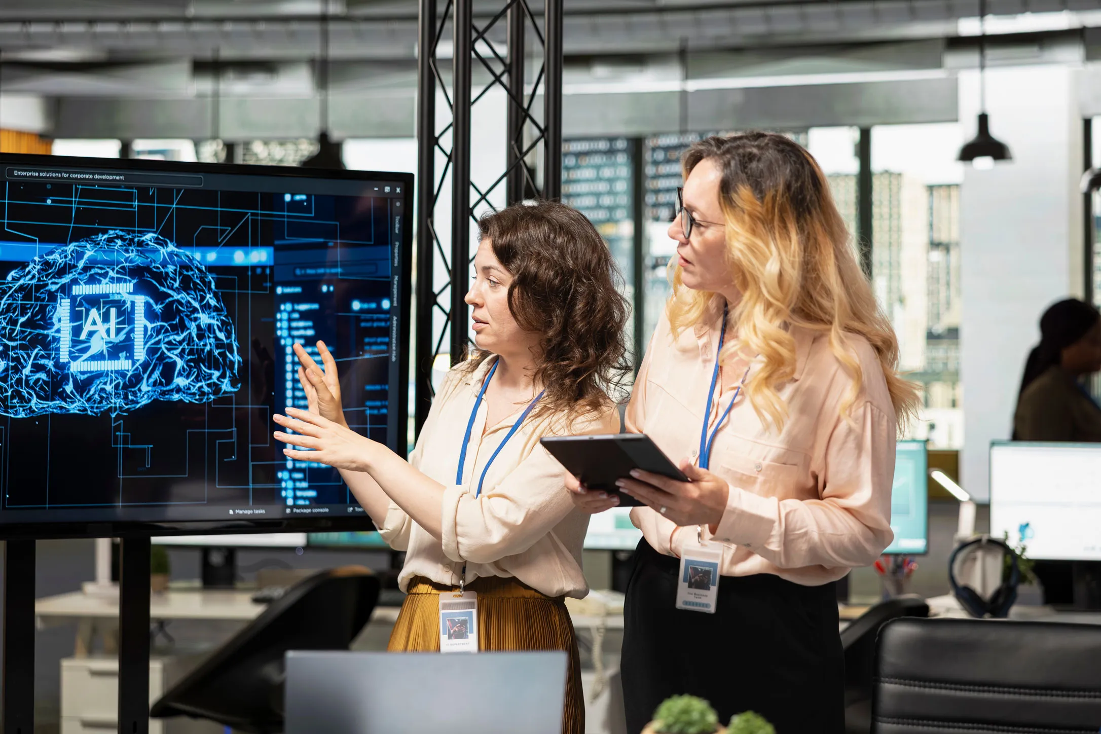 Zwei Frauen besprechen in einem modernen Büro ein digitales Display mit einer Grafik eines Gehirns und dem Schriftzug KI.