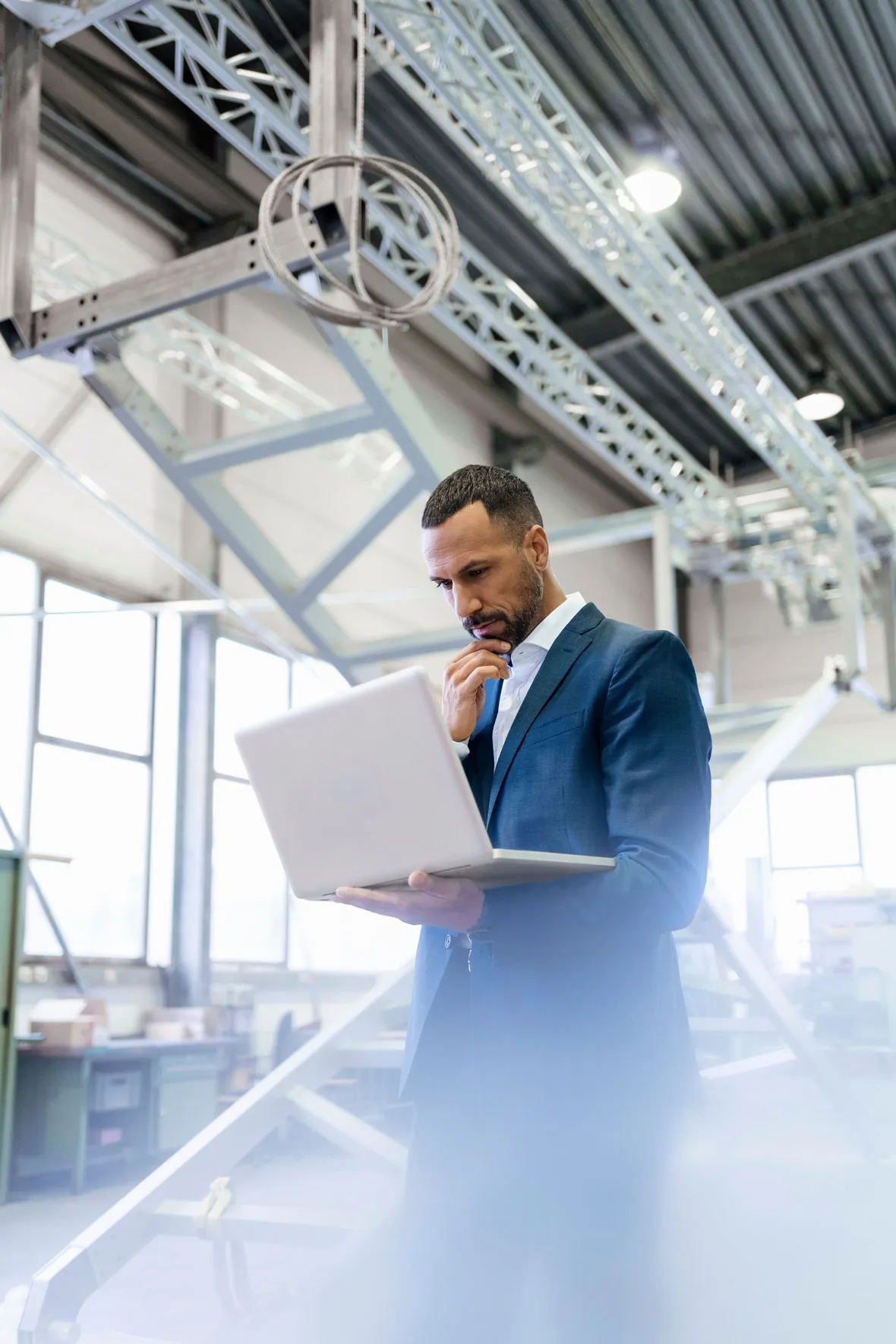 Mann im Anzug steht in einer modernen Industriehalle und schaut nachdenklich auf einen Laptop.