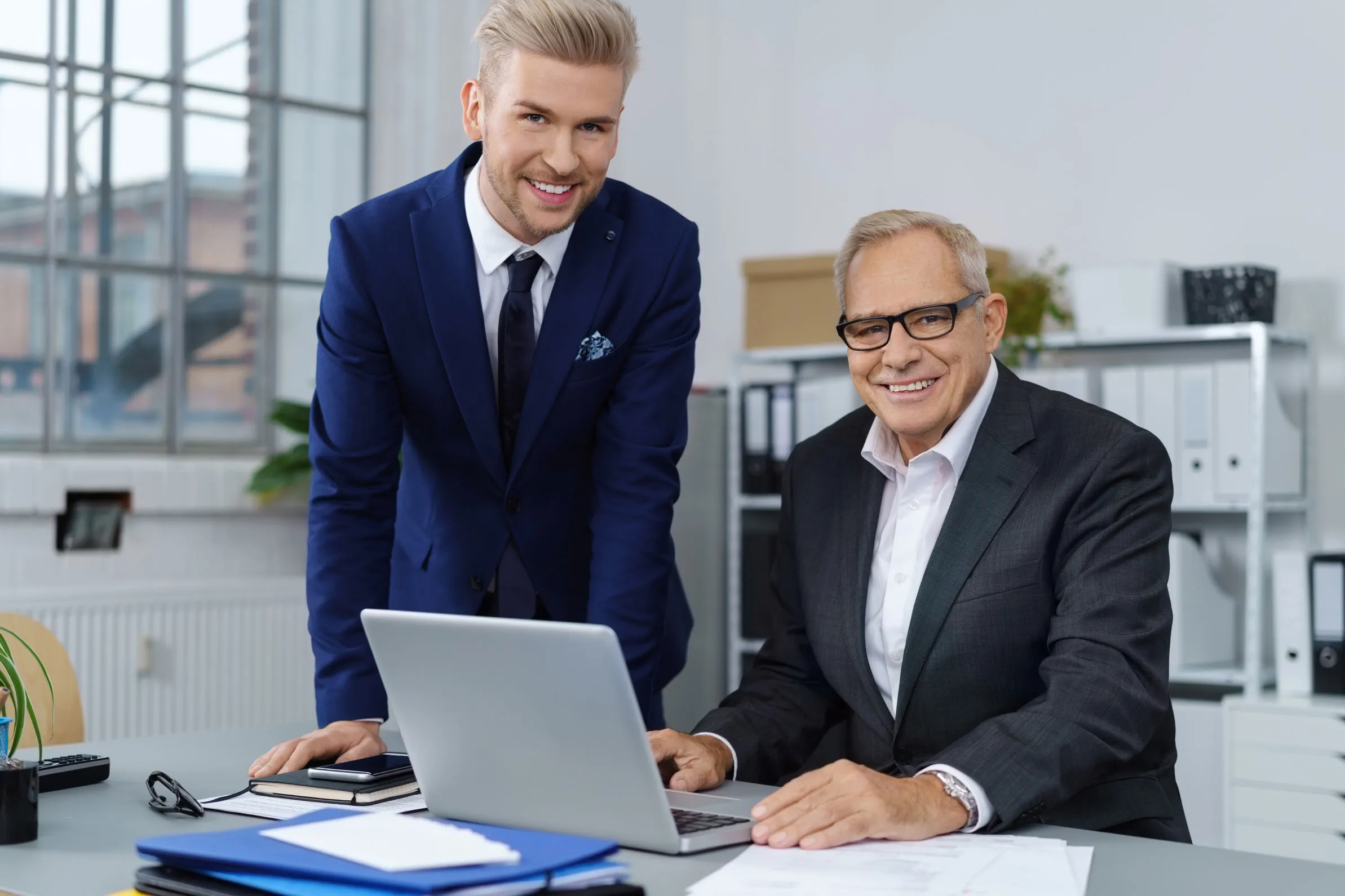 Zwei Geschäftsmänner, eine ältere und eine jüngere, lächeln zusammen an einem Schreibtisch mit Laptop in einem Büro.