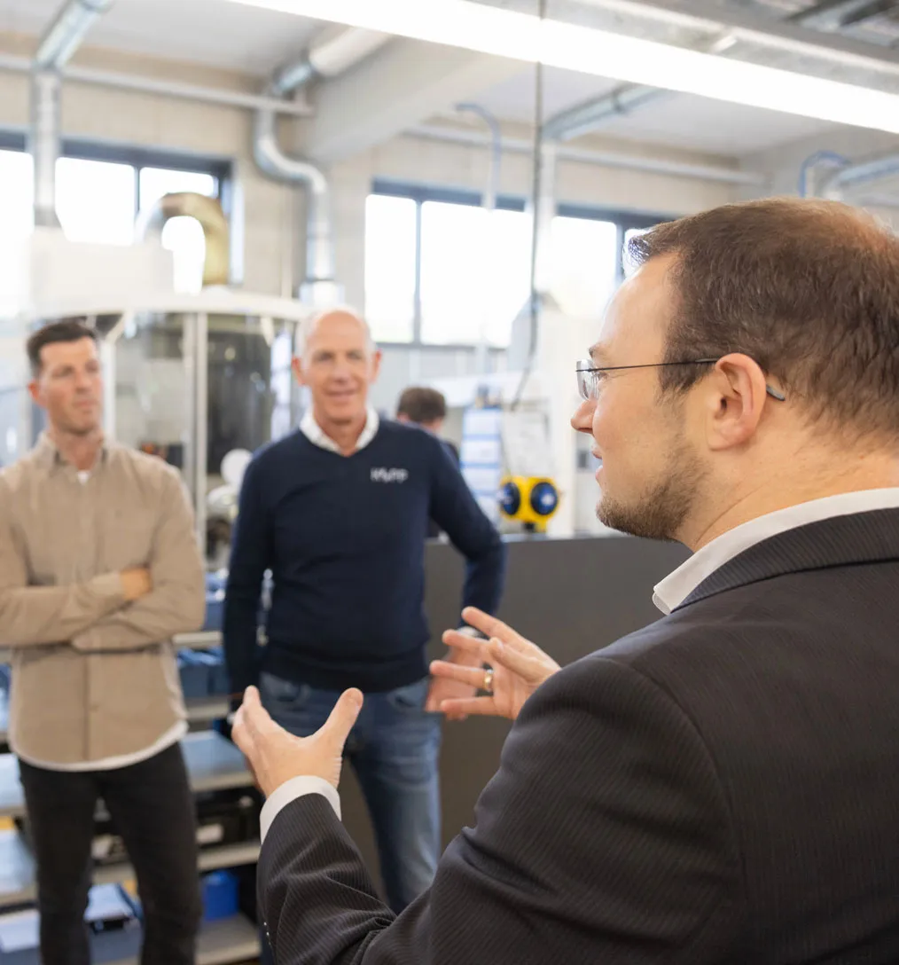 Ein Mann im Anzug erklärt in einer Werkstatt oder Fabrik zwei anderen Männern etwas, die ihm aufmerksam zuhören.