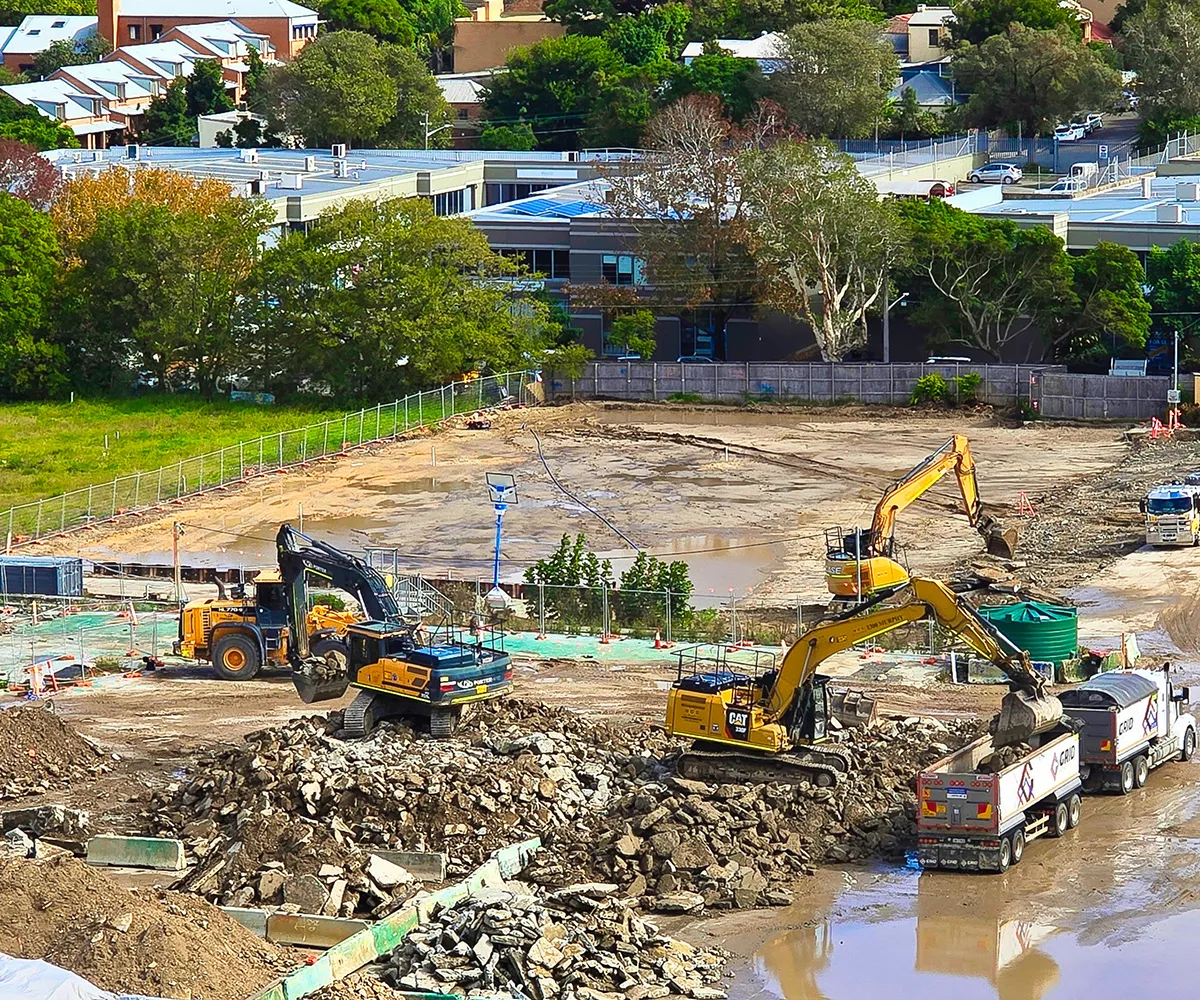 Baustelle mit Baggern, die Gestein auf Muldenkipper laden, umgeben von Gebäuden und Bäumen.