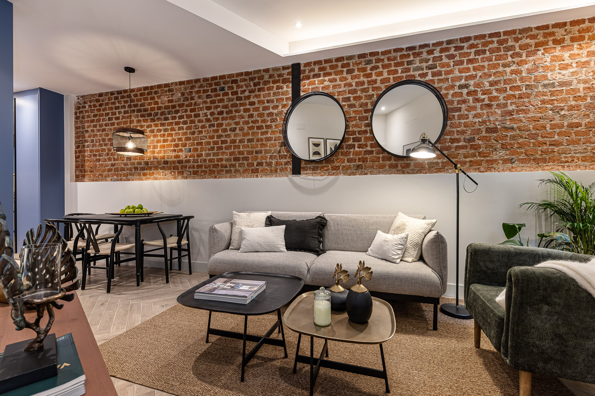 Salón-comedor con muro de ladrillo, sofá gris, mesas bajas y mesa de comedor negra