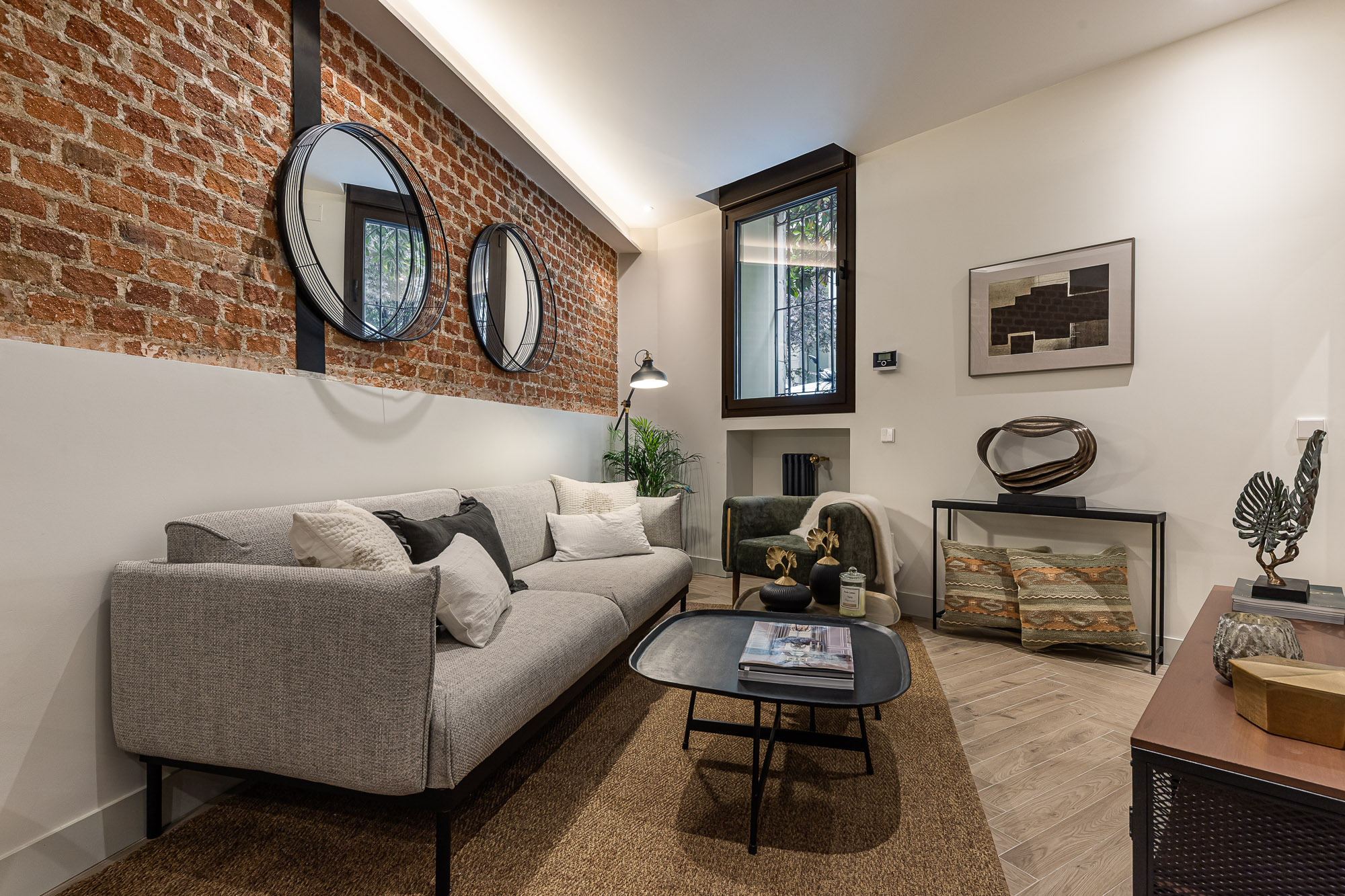 Salón con sofá gris, muro de ladrillo visto a media altura, dos espejos redondos y ventana oscura