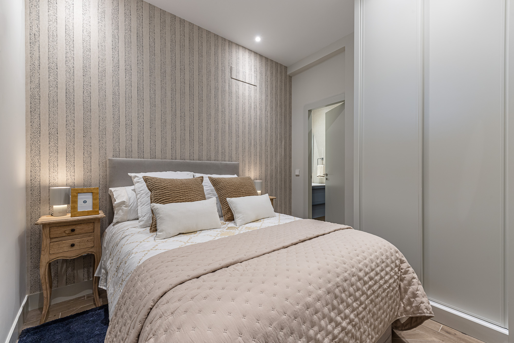 Dormitorio con papel de rayas, cama doble, mesilla de madera y armario empotrado