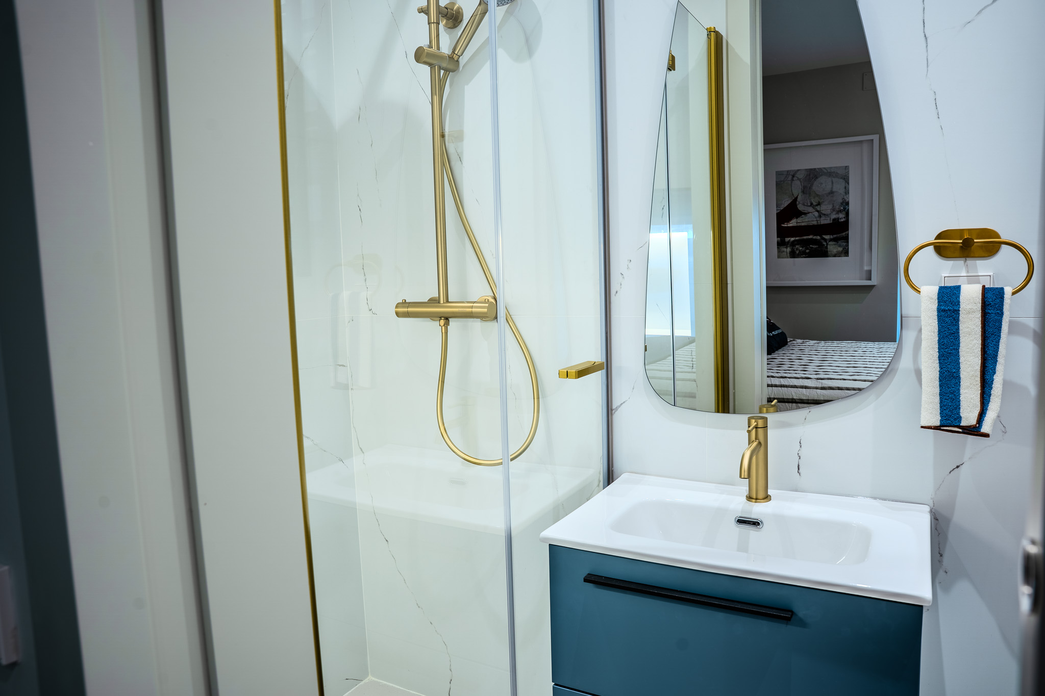 Baño con mueble azul, grifería dorada, espejo orgánico y ducha