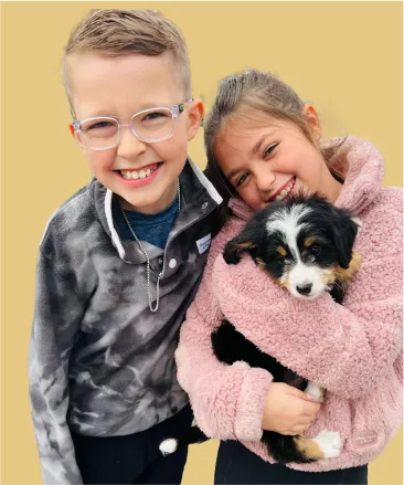 Two smiling children stand close together; the girl in a pink jacket holds a small black, white, and brown puppy.