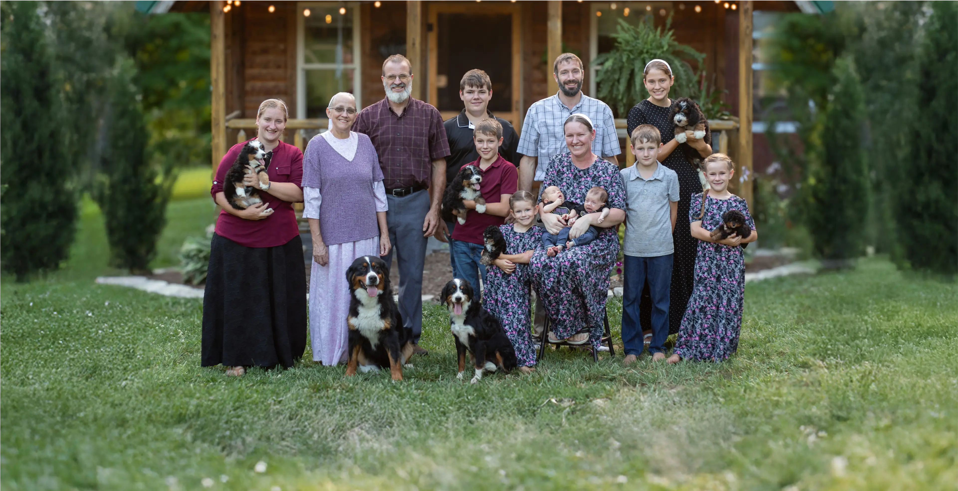 A multi-generational family poses outside a wooden house, with children, adults, several puppies, and two large dogs on a grassy lawn.