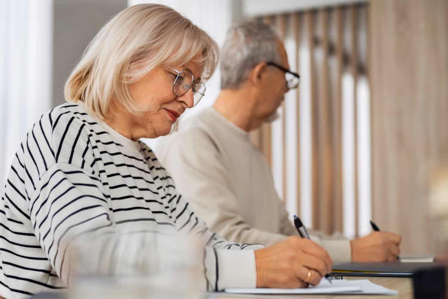 Senior woman and man sitting at a table, writing on paper with pens.