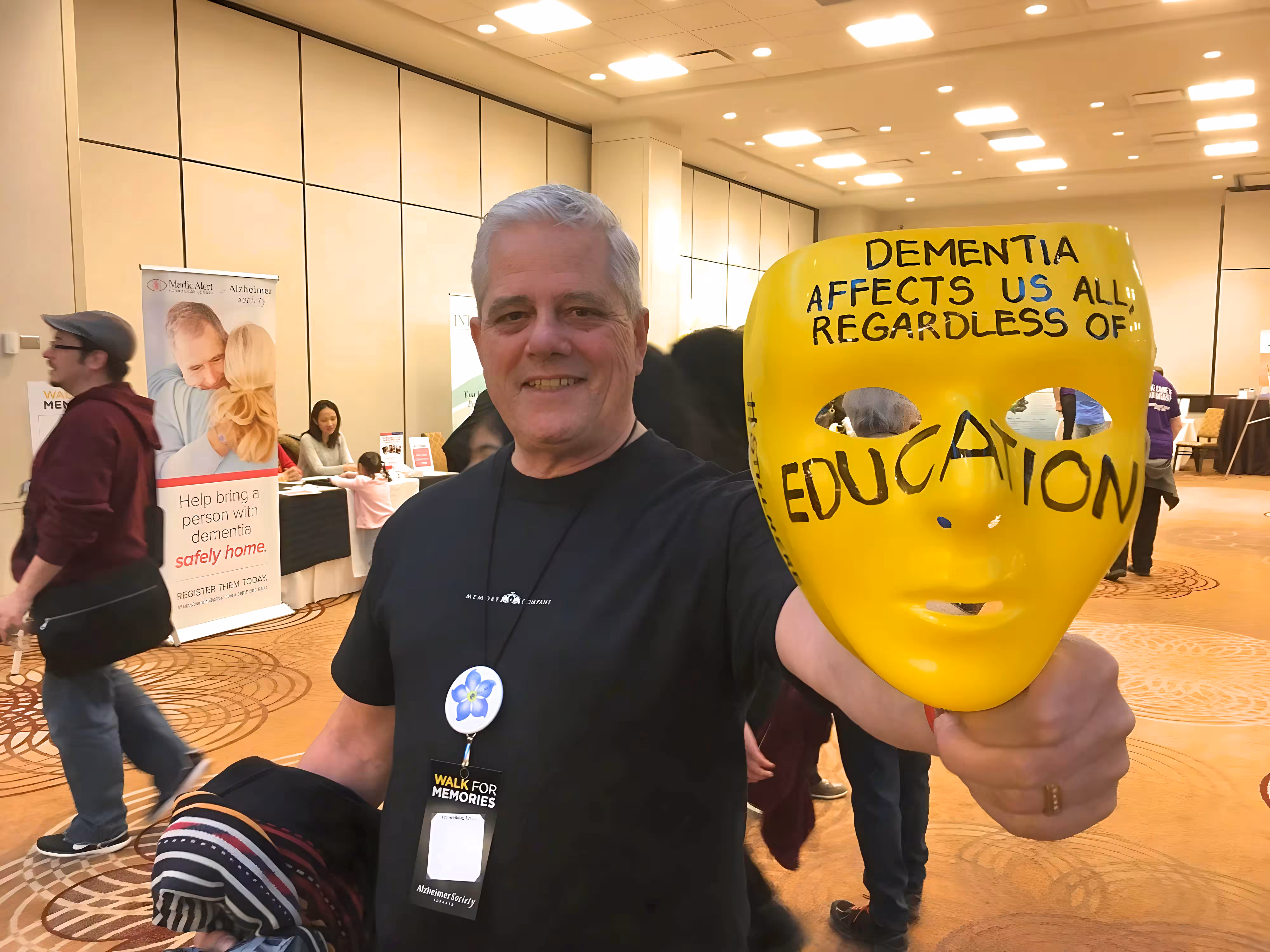 Smiling man at an indoor event holding a yellow mask with the text 'Dementia affects us all regardless of education.'