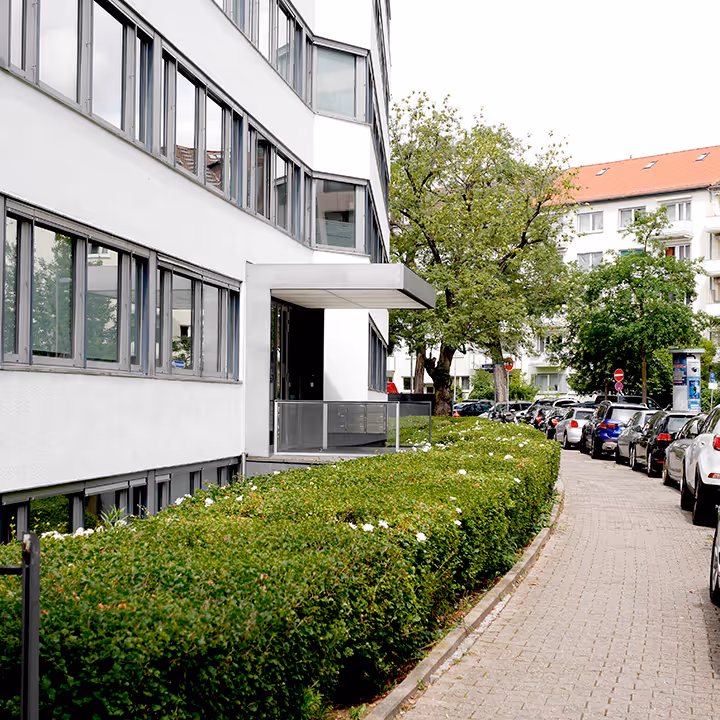 Moderne Wohngebäudefront mit vielen Fenstern, davor blühende grüne Hecken und ein gepflasterter Straßenrand mit geparkten Autos.