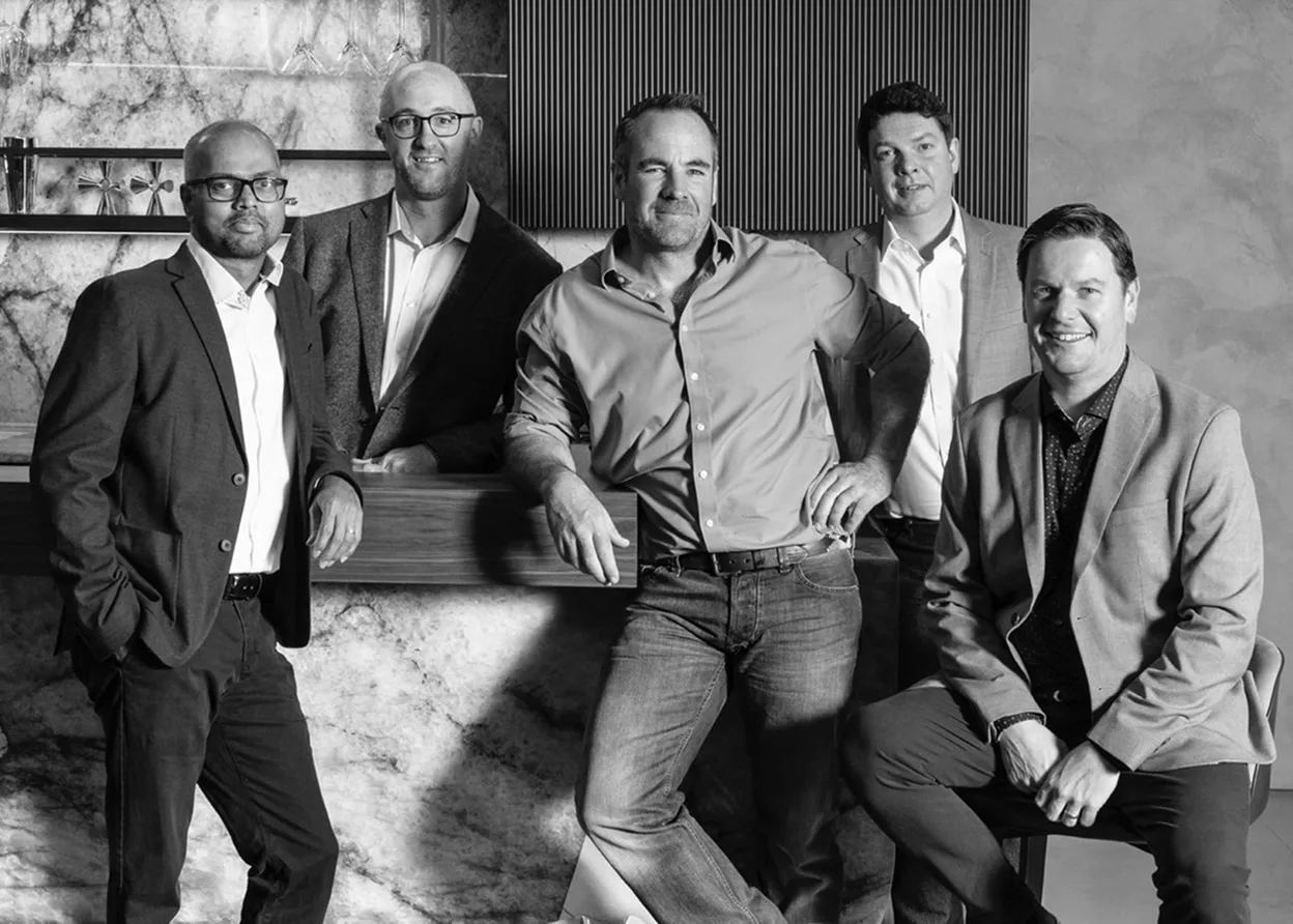 Black and white photo of five men in business casual attire posing in a modern office setting.