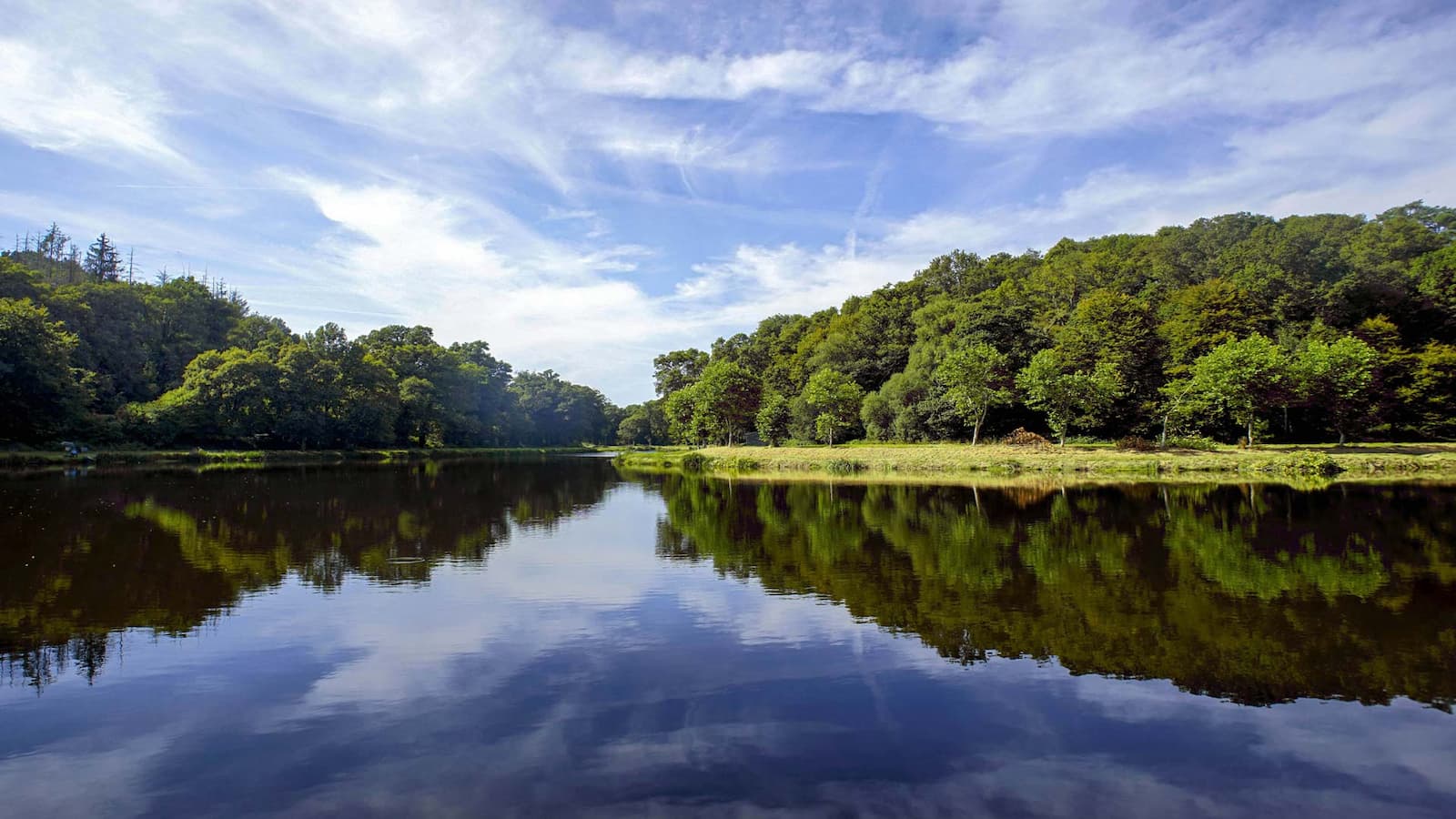 Étang paisible bordé d’une forêt verdoyante sous un ciel bleu en Bretagne, reflétant la nature préservée et le calme des paysages bretons.