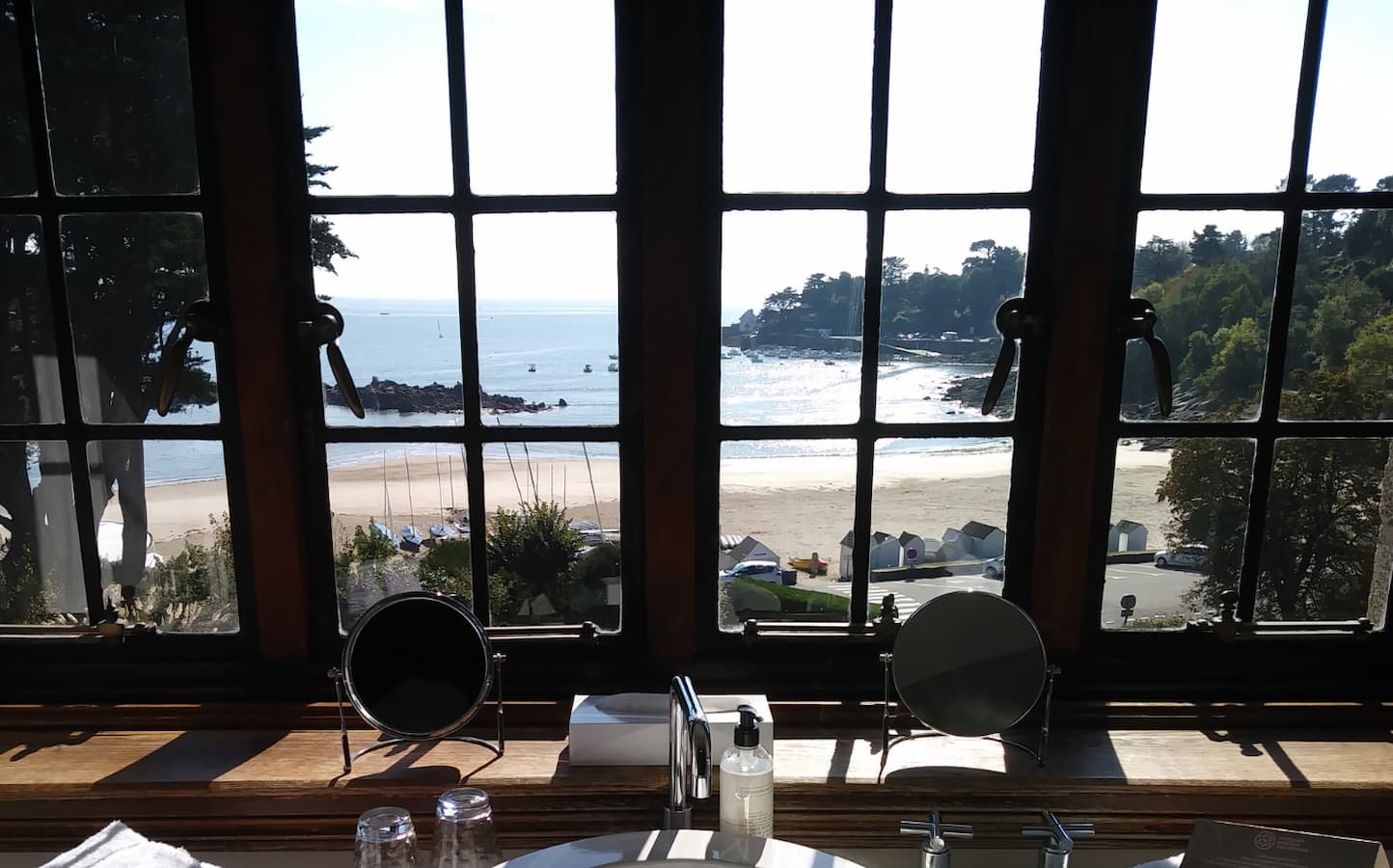 Vue panoramique sur la plage et la mer depuis une salle de bain d’un hôtel de charme en Bretagne, avec fenêtre à carreaux et lumière naturelle.