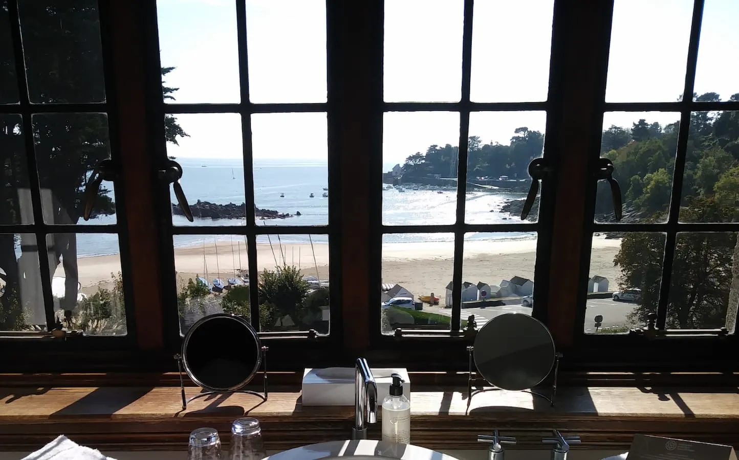 Vue panoramique sur la plage et la mer depuis une salle de bain d’un hôtel de charme en Bretagne, avec fenêtre à carreaux et lumière naturelle.