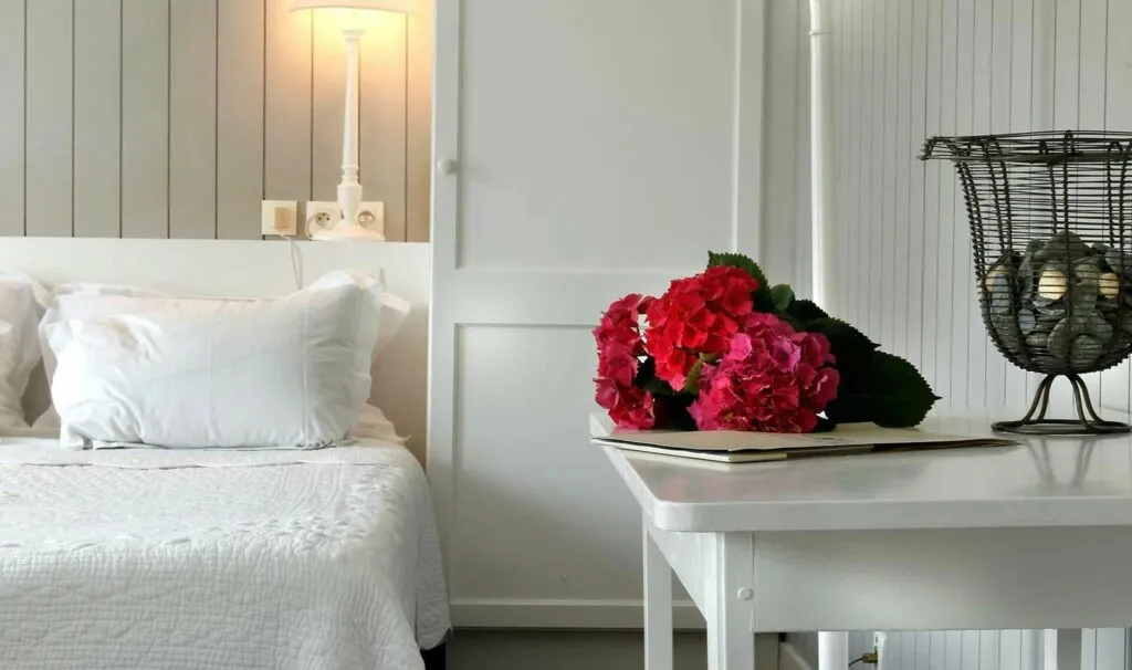 Détail d’une chambre d’hôtel avec lit double, bouquet de fleurs rouges et carnet sur une table blanche, pour un séjour romantique ou en famille en Bretagne