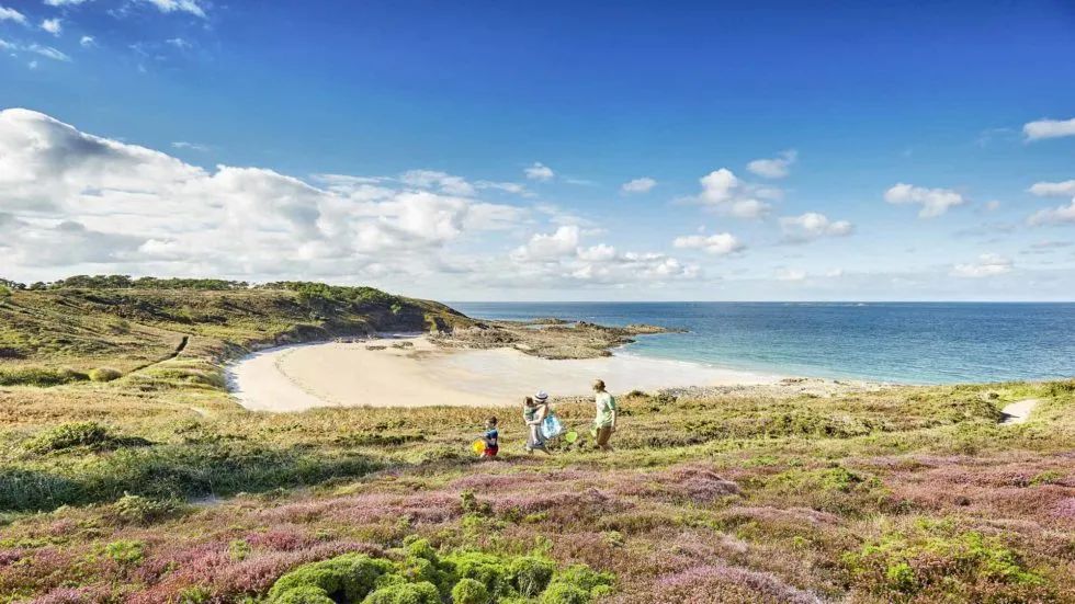 Famille en balade sur un sentier côtier face à une plage sauvage du Golfe du Morbihan, sous un ciel bleu en Bretagne sud