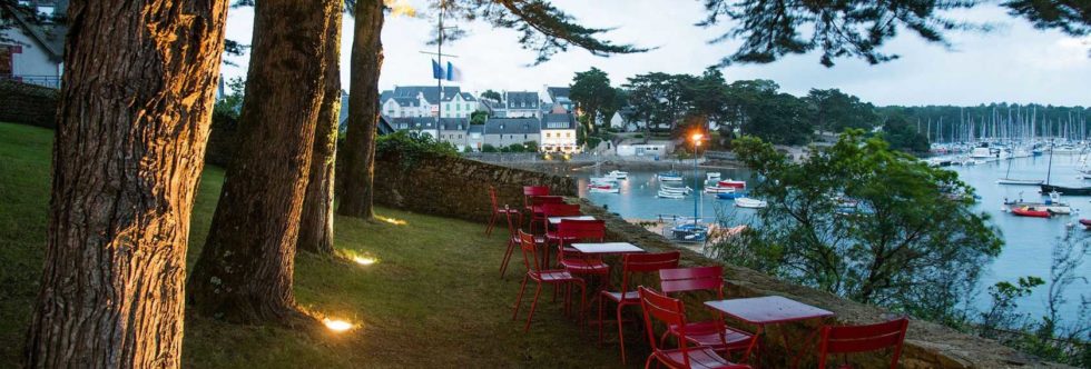Terrace of a hotel-restaurant overlooking a Breton port at dusk, a warm, peaceful atmosphere under the pine trees.