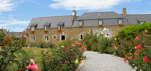Stone facade of La Malouinière des Longchamps, surrounded by a flower garden and paved driveway leading to the entrance.