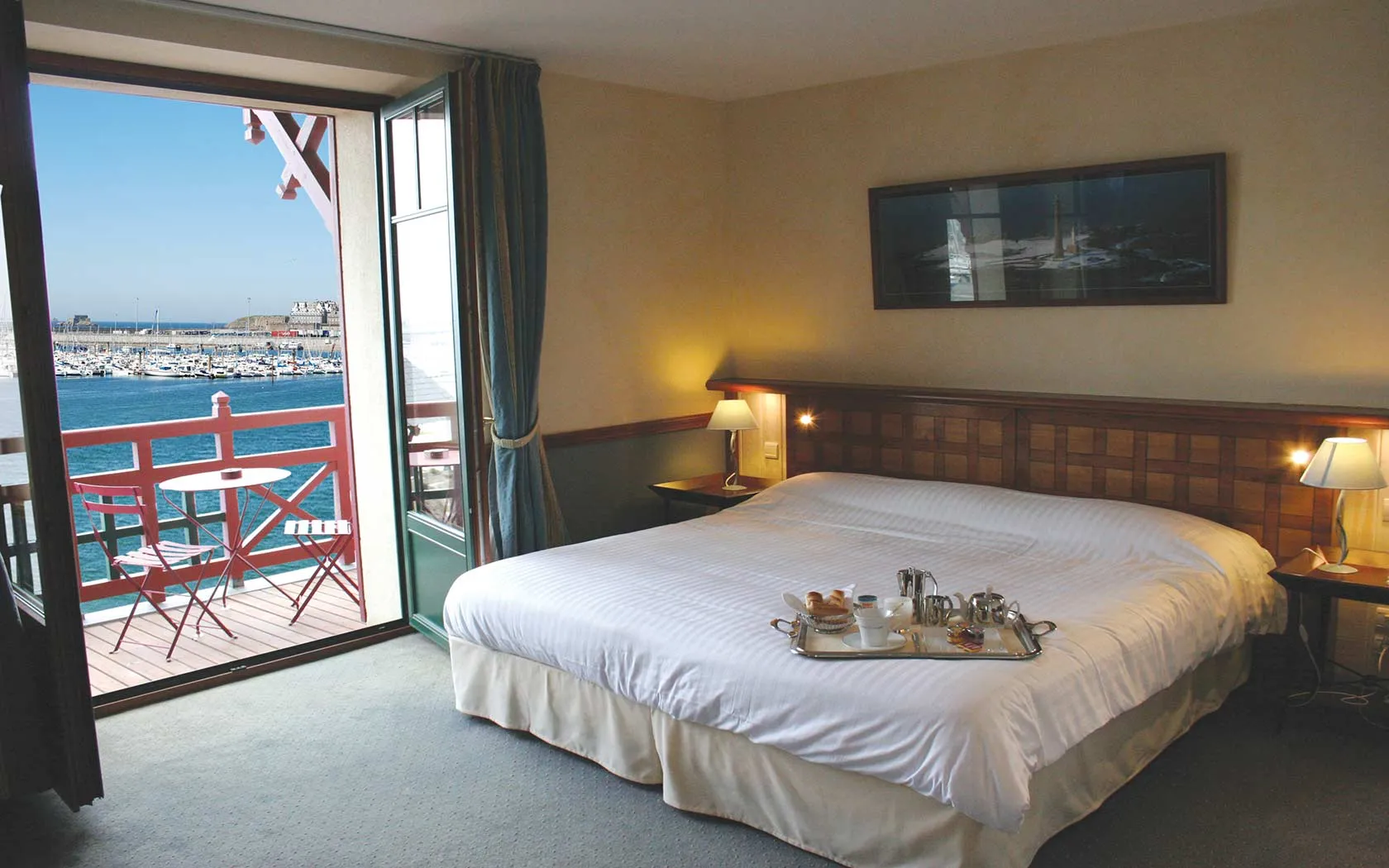 Chambre élégante du Manoir du Cunningham avec lit double, plateau de petit-déjeuner et vue sur le port depuis le balcon.
