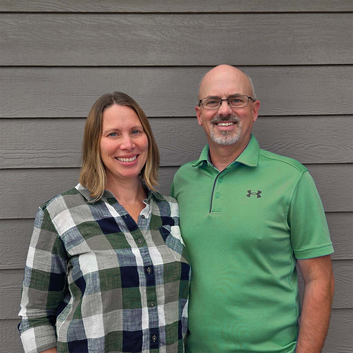 Smiling woman in plaid shirt and man in green polo standing against gray siding