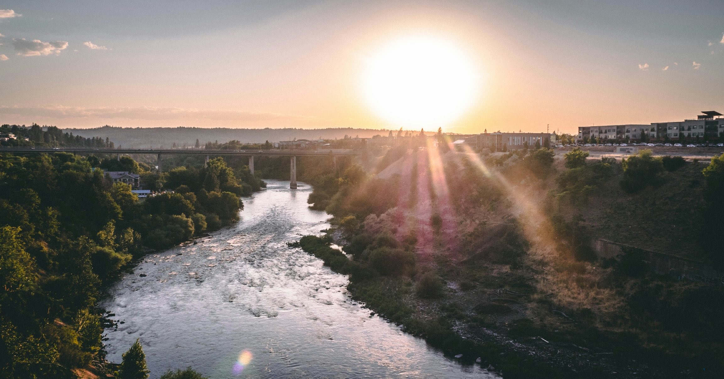 Sunset view over the Spokane River with a bridge in the distance, surrounded by trees, hills, and buildings along the riverbanks.
