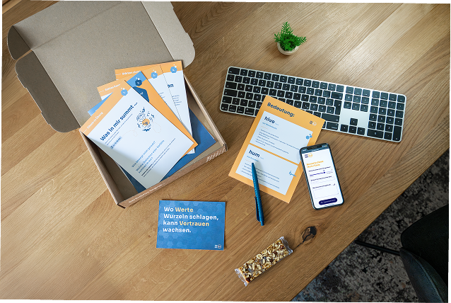 Ein Arbeitsplatz mit einer Tastatur, einer Pflanze, einem Smartphone, Karteikarten mit deutschen Texten, einem blauen Kärtchen mit dem Spruch 'Wo Werte Wurzeln schlagen, kann Vertrauen wachsen.' und einem Müsliriegel auf einem Holztisch.