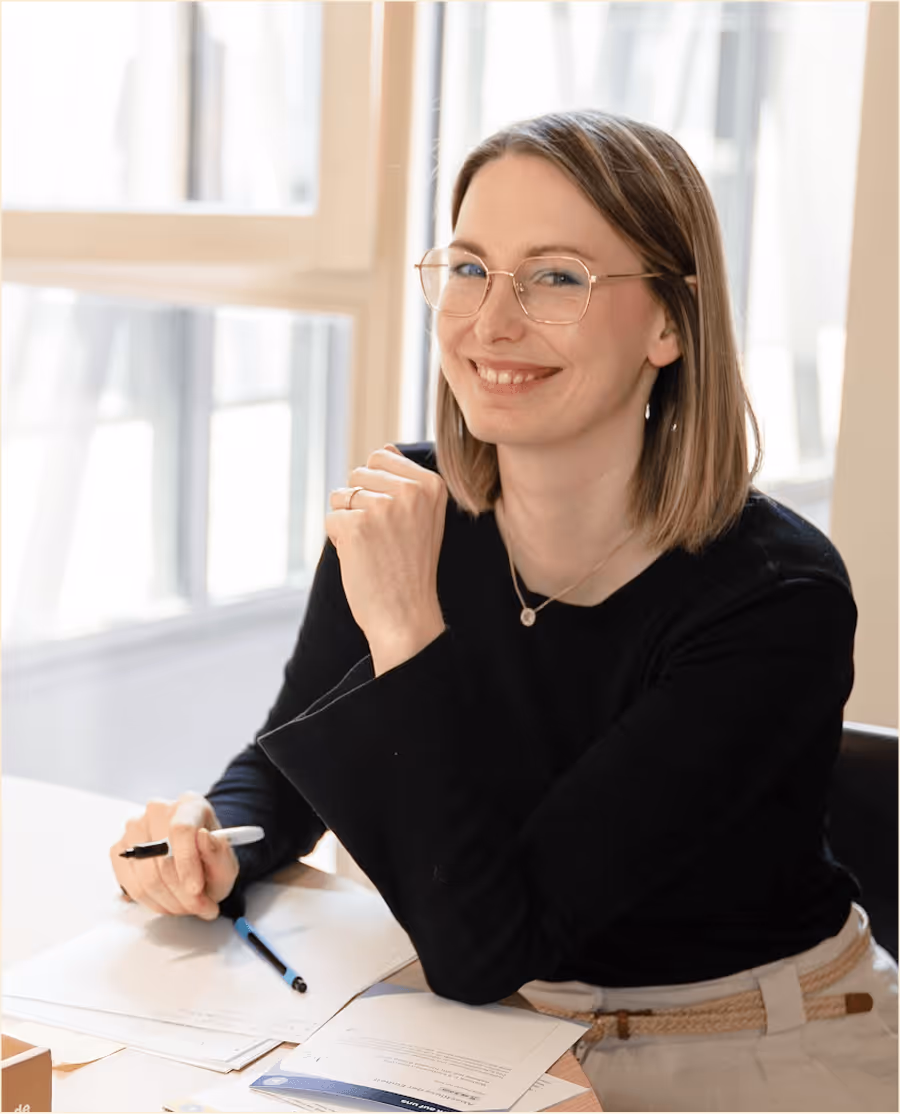 Lächelnde Frau mit Brille sitzt an einem Tisch mit geöffnetem Notizbuch und Dokumenten, hält Stift in der Hand.