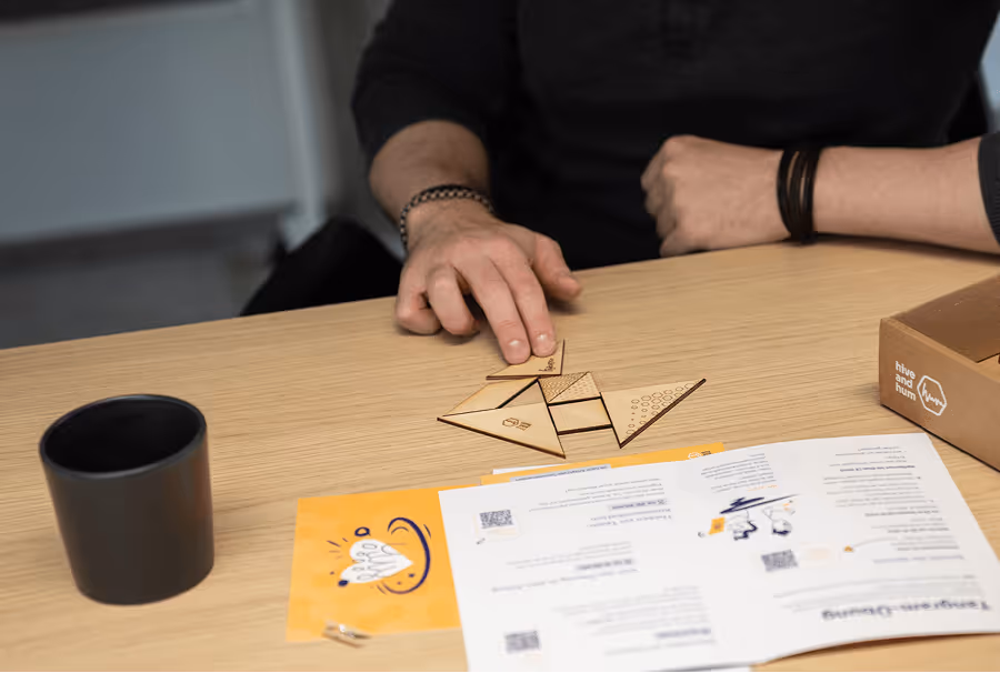 Eine Person arrangiert Holzteile eines Tangram-Puzzles auf einem Holztisch neben Anleitung und einer schwarzen Tasse.