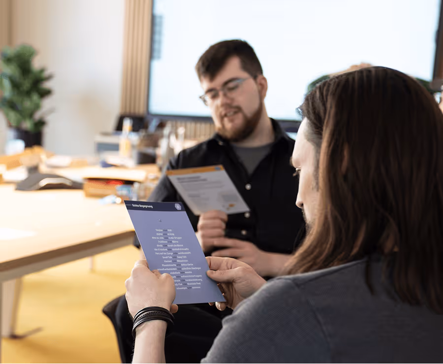 Zwei Männer in einem Büro lesen Informationsblätter, der eine im Vordergrund mit langen Haaren, der andere im Hintergrund mit Brille und Bart.