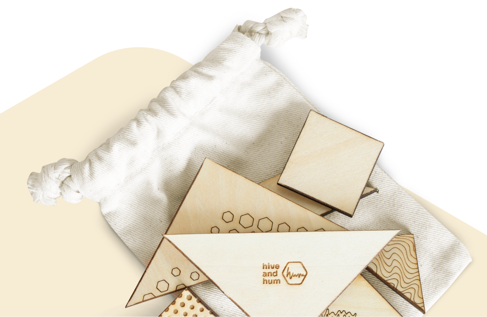 Weiße Stofftasche mit flachen, holzfarbenen geometrischen Puzzleteilen mit Mustern und dem Schriftzug 'hive and hum'.