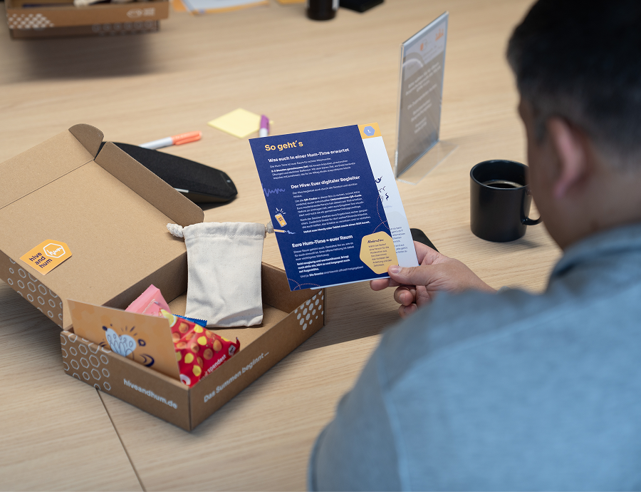 Ein Mann sitzt an einem Holztisch und liest eine blaue Informationskarte aus einem geöffneten Karton mit verschiedenen kleinen Gegenständen.