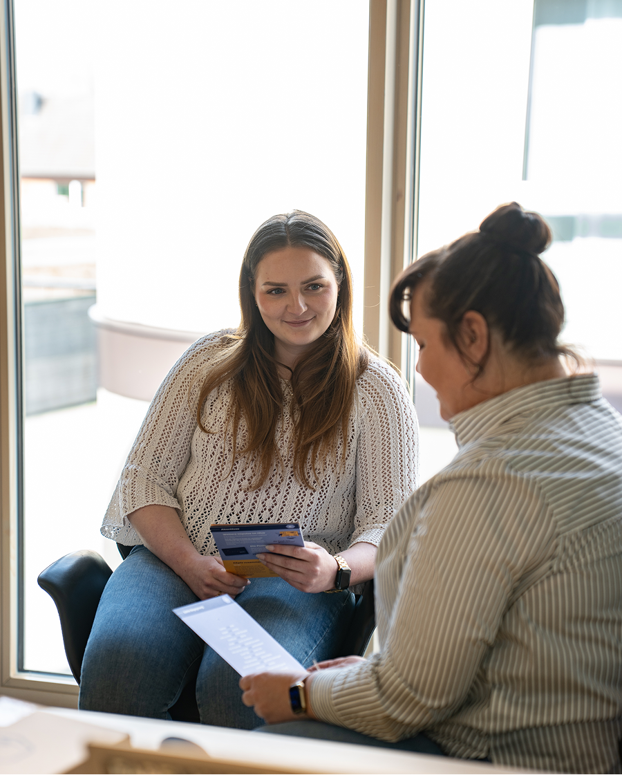 Zwei Frauen sitzen in einem hellen Raum und führen ein Gespräch, während sie Informationsbroschüren halten.