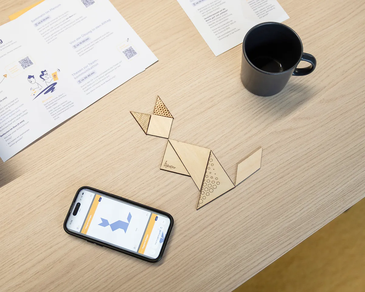 Ein Holz-Tangram-Puzzle auf einem Holztisch, geformt wie eine Katze, neben einem Smartphone mit Tangram-Anleitung und einer schwarzen Kaffeetasse.