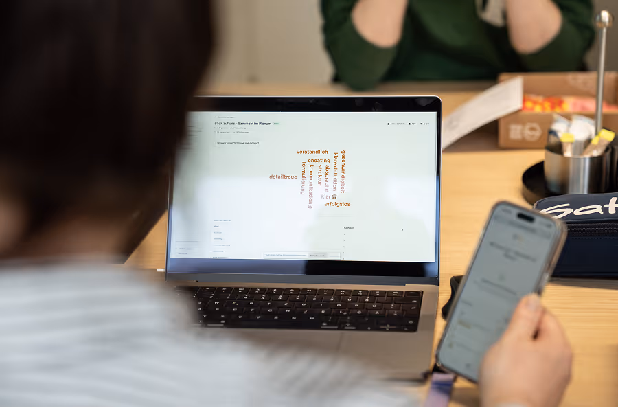 Person sieht auf Laptopbildschirm mit Wortwolke und hält ein Smartphone in der Hand.