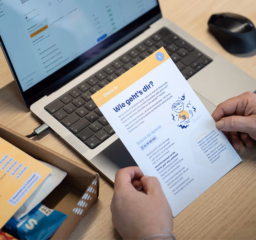 Person hält ein Merkblatt auf Deutsch mit dem Titel 'Wie geht's dir?' vor einem Laptop und einer schwarzen Maus auf einem Holztisch.