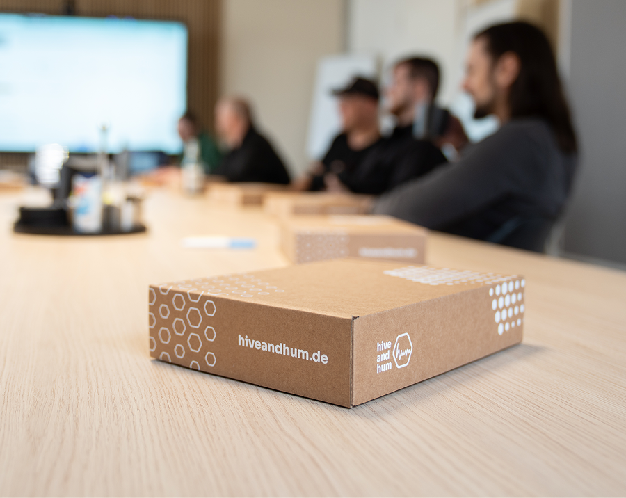 Braune Versandbox mit Aufdruck 'hiveandhum.de' auf einem Konferenztisch, im unscharfen Hintergrund sitzen mehrere Menschen.