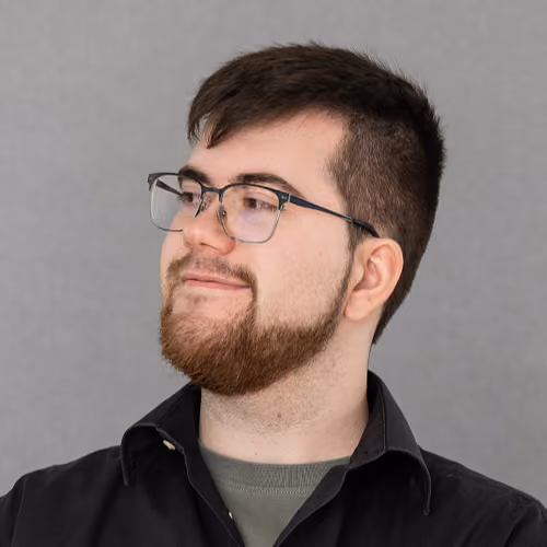 Junger Mann mit braunem Bart und Brille blickt lächelnd seitlich, trägt schwarzen Oberteil und olivgrünes T-Shirt.