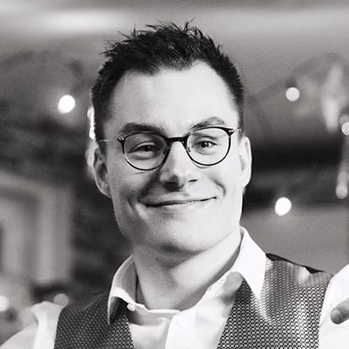 Lächelnder Mann mit dunkler Kurzhaarfrisur, Brille und Weste in einem unscharfen Innenraum.