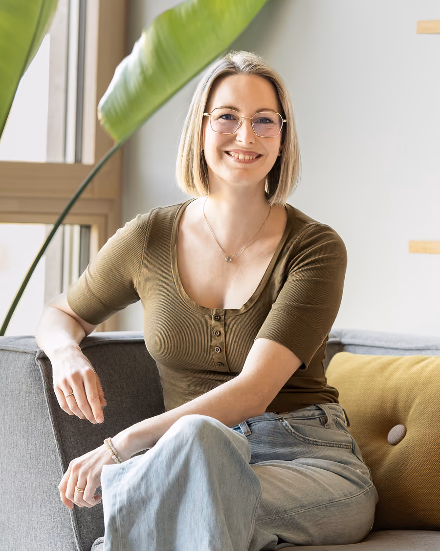 Frau mit kurzem blondem Haar, Brille und olivgrünem Oberteil, die auf einem Sofa sitzt und lächelt.