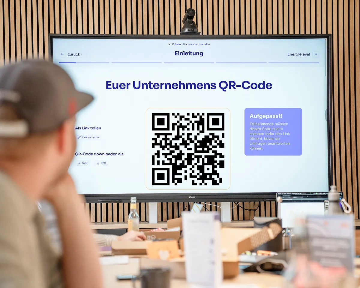 Person schaut auf großen Bildschirm mit Unternehmens-QR-Code, Text und Anweisungen zum Teilen und Herunterladen des QR-Codes in SVG- und JPG-Formaten, sowie einer Warnung, dass Teilnehmer den Code zuerst scannen müssen, um Umfragen zu beantworten.