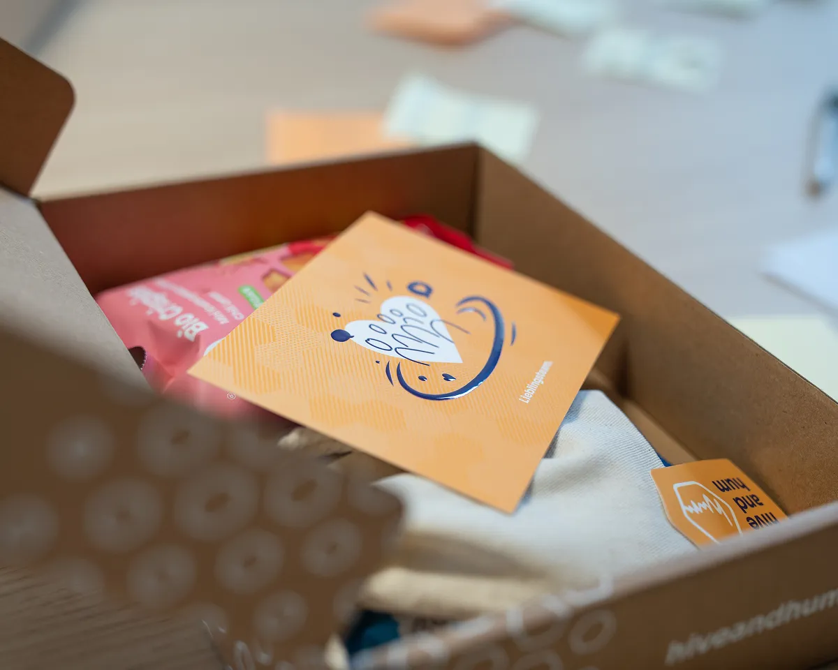 Offener Karton mit Taschentuch, orangefarbener Karte und Snackverpackung auf einem Tisch.