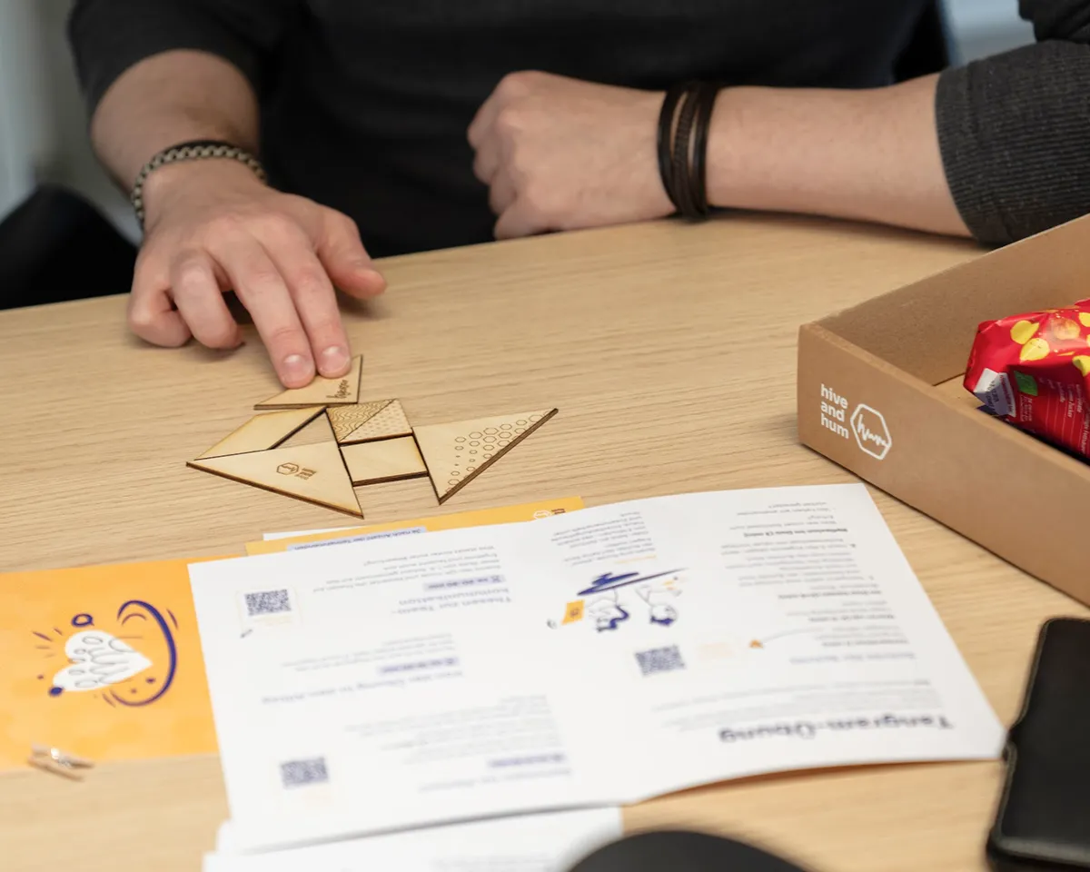 Eine Person arrangiert Holzteile eines Tangram-Puzzles auf einem Holztisch neben Anleitung und einer schwarzen Tasse.