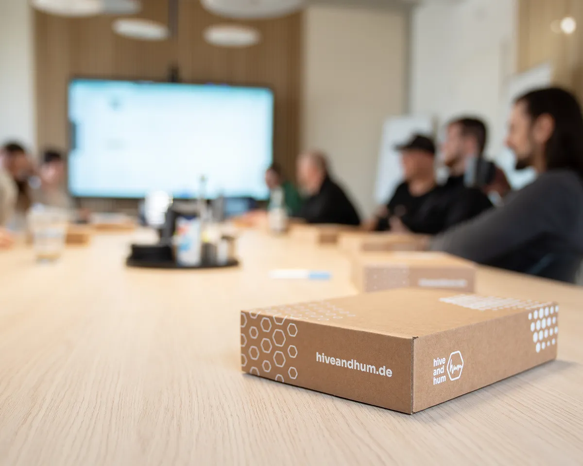 Braune Versandbox mit Aufdruck 'hiveandhum.de' auf einem Konferenztisch, im unscharfen Hintergrund sitzen mehrere Menschen.