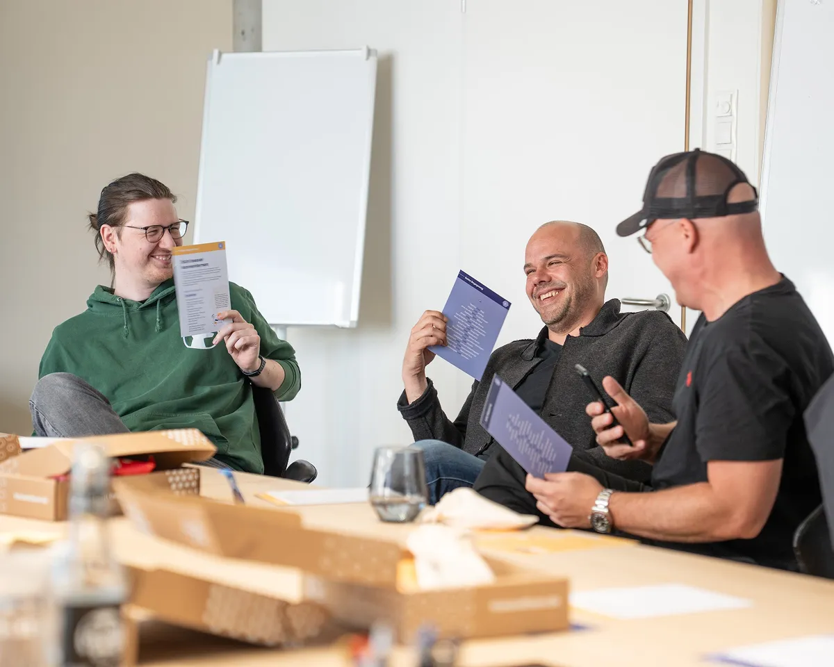 Drei Männer sitzen an einem Tisch, lachen und halten gedruckte Dokumente in einem Besprechungsraum.