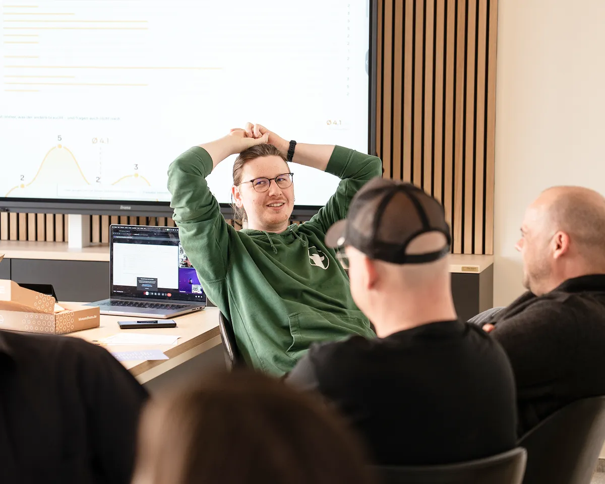 Drei Männer sitzen in einem Meetingraum mit Laptop und Diagrammen auf einem großen Bildschirm im Hintergrund.
