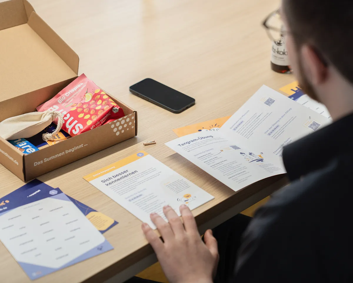 Person sitzt an einem Holztisch und liest Informationsblätter, daneben eine offene Box mit Snacks und ein Smartphone.