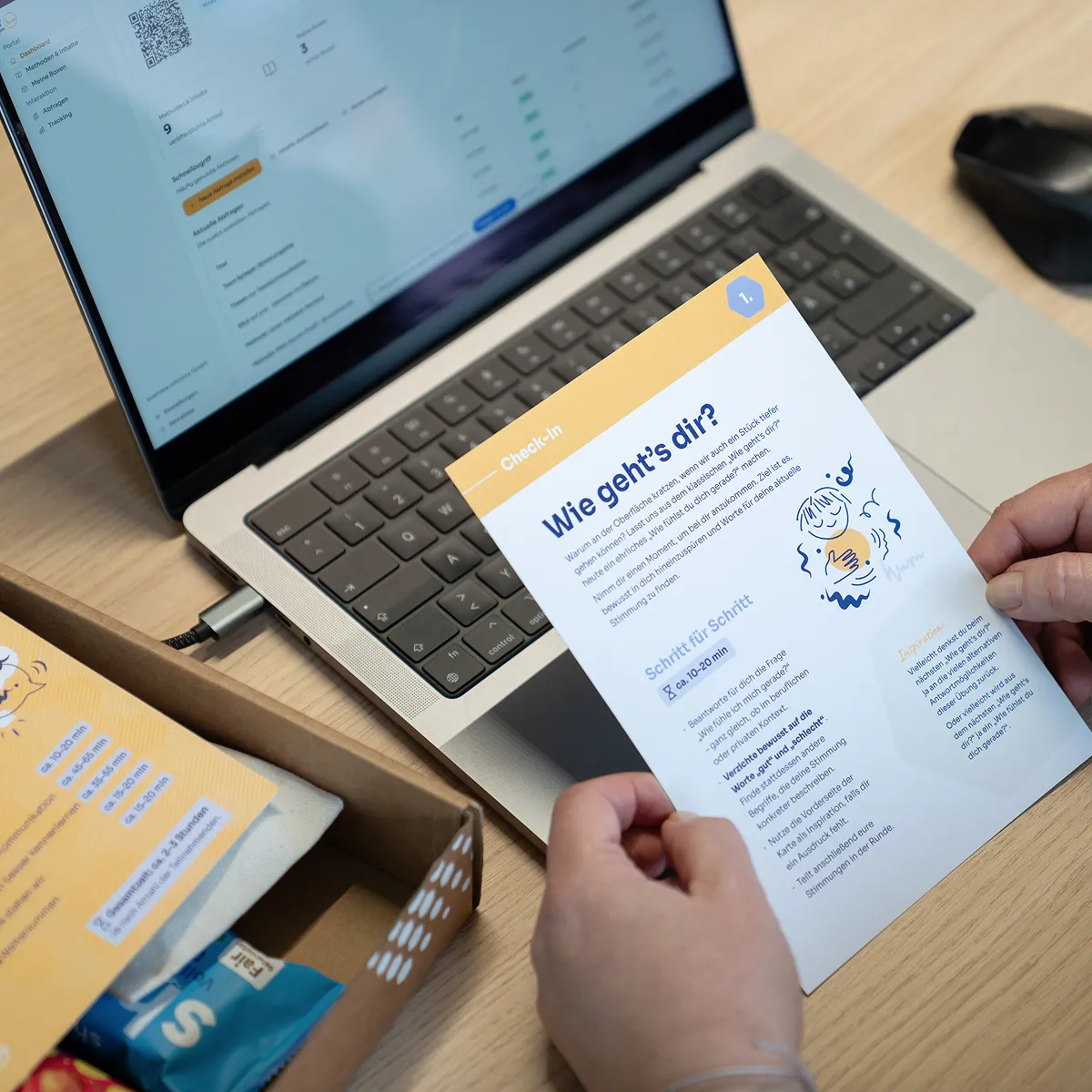 Person hält ein Merkblatt auf Deutsch mit dem Titel 'Wie geht's dir?' vor einem Laptop und einer schwarzen Maus auf einem Holztisch.