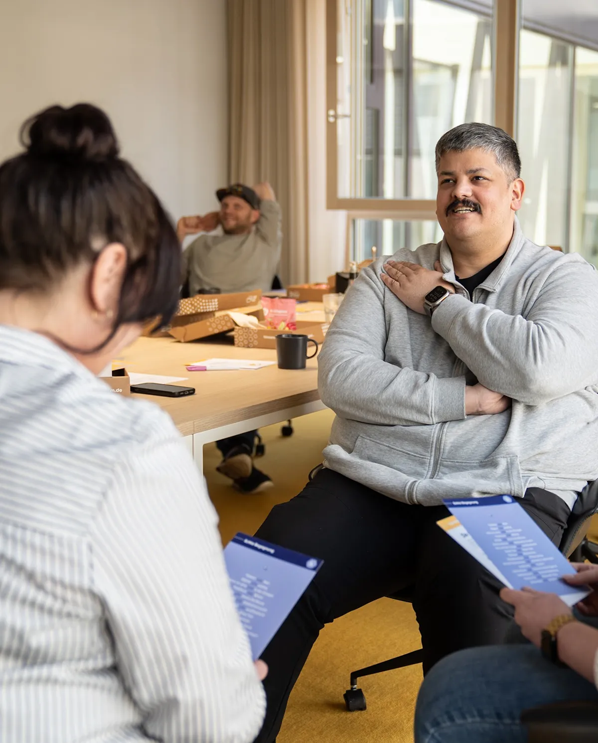 Drei Erwachsene sitzen im Büro, zwei Frauen mit Dokumenten auf dem Schoß und ein Mann in grauem Pulli mittig, der spricht.