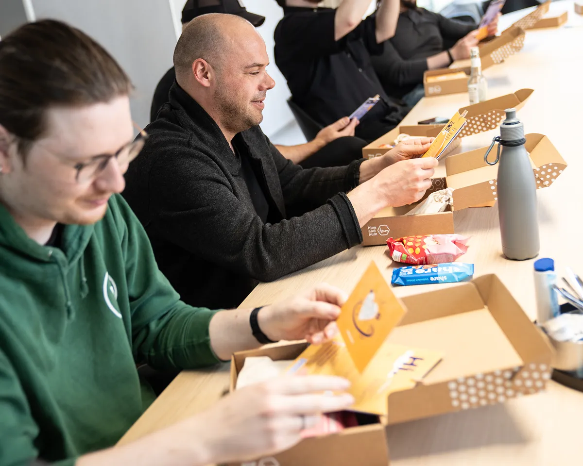 Mehrere Personen sitzen an einem Tisch und öffnen braune Pappschachteln, in denen Snacks und Unterlagen enthalten sind.