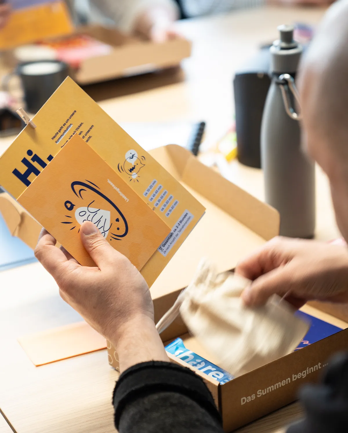 Person öffnet eine braune Schachtel mit Aufdruck "Das Summen beginnt ..." und hält ein gelbes Heft und eine Karte mit einer Zeichnung von einem summenden Insekt in der Hand, auf dem Tisch eine Trinkflasche und weitere Gegenstände.